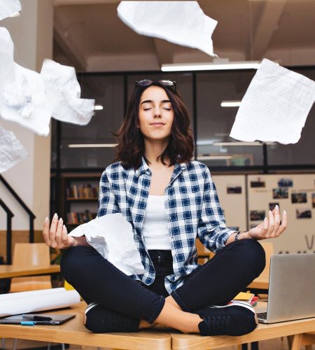 Young pretty joyful brunette woman meditating on table surround work stuff and flying papers. Cheerful mood, taking a break, working, studying, relaxation, true emotions.