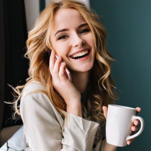 young woman talking phone and laughing with cup of coffee tea in hand happy morning she has beautiful wavy blonde hair room with blue turquoise wall wearing nice lace pajama