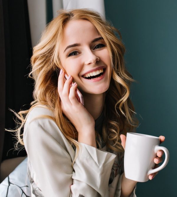 young woman talking phone and laughing with cup of coffee tea in hand happy morning she has beautiful wavy blonde hair room with blue turquoise wall wearing nice lace pajama e1627362630940