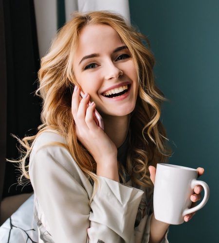 Young woman talking phone and laughing with cup of coffee, tea in hand, happy morning. She has beautiful wavy blonde hair. Room with blue, turquoise wall on background. Wearing nice lace pajama..